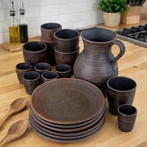 May include: A rustic brown ceramic dinnerware set, including plates, cups, a pitcher, and small bowls. The set is arranged on a wooden surface, with wooden spoons visible in the foreground. The pottery has a textured finish.
