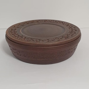 Puede incluir: Una caja redonda de madera marrón con tapa. La tapa y la base de la caja presentan intrincados diseños florales tallados. La caja es de color marrón medio cálido y se encuentra sobre una superficie blanca.