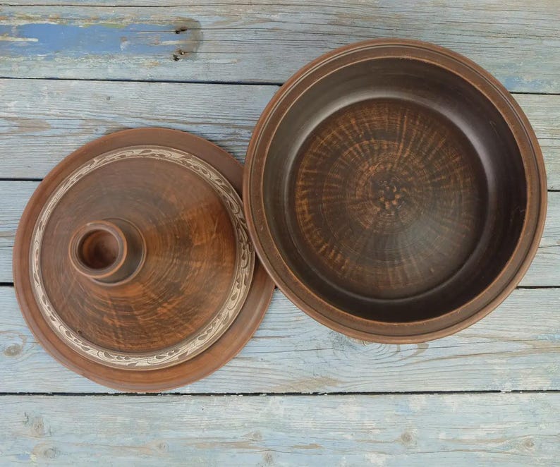 May include: A brown ceramic tagine pot with a lid, set against a weathered blue wooden surface. The lid features a central knob and a decorative border. The pot's interior is visible, showcasing its depth and texture.