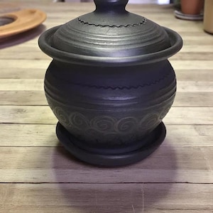 Op de afbeelding: Een donkergrijze keramische pot met een deksel, op een bijpassende schotel. De pot heeft een afgeronde vorm met geëtste horizontale lijnen en een decoratief wervelpatroon. Het deksel heeft een knop en een geschulpte rand.