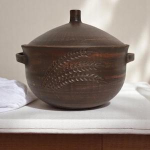 Peut inclure: Une marmite de cuisine en céramique marron avec un couvercle. Le pot a une forme arrondie avec deux poignées et un motif décoratif d'épis de blé. Le couvercle a un petit bouton. Le pot est posé sur une surface blanche.