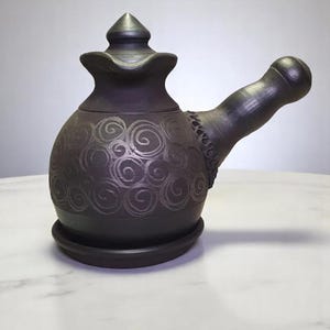 Könnte beinhalten: Eine dunkelbraune Keramik-Teekanne mit einem dekorativen Wirbelmuster. Die Teekanne hat einen abgerundeten Körper, einen gebogenen Griff und einen Deckel mit einer spitzen Spitze. Die Teekanne steht auf einer passenden Untertasse.