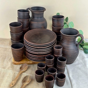 May include: A rustic, handmade pottery dinnerware set in shades of brown. The set includes plates, cups, pitchers, and wooden spoons. The plates are stacked, and the cups are arranged in neat rows. The pitchers have handles.