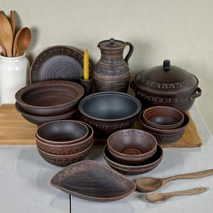 May include: A selection of brown ceramic kitchenware, including bowls, plates, a jug, and a lidded pot. Some pieces feature decorative floral patterns around the rims. Wooden spoons and a candle holder with a yellow candle are also visible.