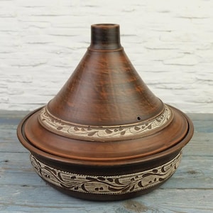 Op de afbeelding: Een bruine keramische tajinepot met een conisch deksel en een ronde basis. De pot heeft een decoratieve band met witte bloemmotieven rond het deksel en de basis. De tajine is ontworpen voor het koken en serveren van Marokkaanse gerechten.