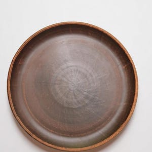 Puede incluir: Un plato redondo de cerámica marrón con un borde ligeramente elevado. El plato tiene un tono oscuro y terroso con patrones de vetas de madera visibles. El borde es de un tono marrón más claro, creando un contraste sutil. Ideal para servir alimentos.