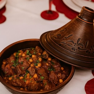 以下が含まれることがあります： 伝統的なモロッコ風タジン料理。濃い茶色の陶器の鍋に円錐形の蓋がついています。料理には煮込んだ肉、ひよこ豆、ハーブが入っています。テーブルセッティングには赤いアクセントとワイングラスが含まれています。