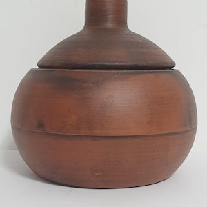 以下が含まれることがあります： 蓋付きの茶色の陶器の壺。壺は丸みを帯びた底と円筒形の首を持っています。蓋はわずかにドーム型で、壺はテラコッタまたは同様の素材で作られているようです。壺は保管に使用される可能性があります。寸法はセンチメートルです。