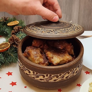 Może przedstawiać: Brązowe ceramiczne naczynie do zapiekania z pokrywką, wypełnione ugotowanym jedzeniem. Naczynie ma dekoracyjny wzór na obrzeżu i na pokrywce. Jedzenie w środku wydaje się być pieczonym kurczakiem lub mięsem. Naczynie stoi na stole z uroczystym obrusem.