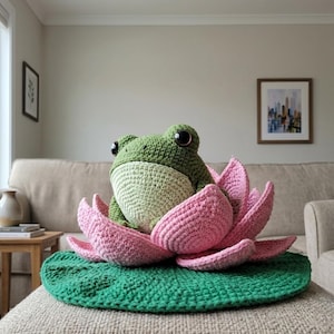 May include: A crocheted green frog with large dark eyes and a light cream-colored belly, sitting inside a large pink lotus flower. The flower rests on a flat, circular green lily pad, all crafted from yarn with a textured, handmade appearance.
