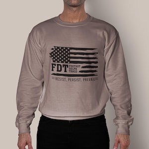 May include: A tan long-sleeve sweatshirt with a black and white American flag graphic. The text "FDT Fox Trot Delta Tango Resist, Persist, Prevail" is printed below the flag.