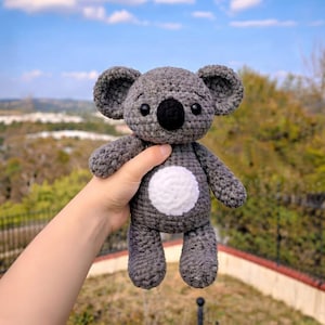 May include: A handmade gray crochet koala bear with a white belly patch, black eyes, and a black nose. The koala has large, rounded ears and is held up against a blurred outdoor background.
