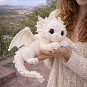 May include: A crocheted white dragon with large black eyes, blush-pink cheeks, and small wings. The dragon is lying down, with its paws and tail curled. The dragon is a handmade item.