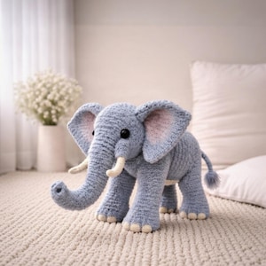 May include: A handmade, light blue crochet elephant with large ears, a trunk, and small tusks. The elephant has black eyes and a small tail. The ears have a pink interior. The elephant is standing on a cream-colored rug.