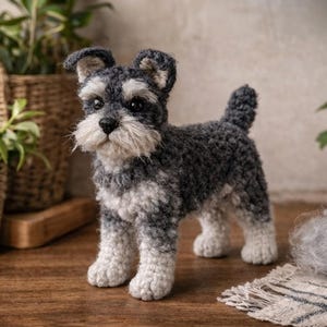 May include: A handmade crochet Schnauzer dog figurine in shades of gray and white. The dog has a fluffy beard and eyebrows, with a small tail and perky ears. The dog is standing on a wooden surface.