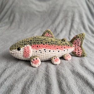 May include: A crocheted stuffed fish toy featuring a dark olive green back with black spots, a white underbelly, and a prominent pink horizontal stripe along the side. The fins and tail are a mix of olive green and pink with black speckles.
