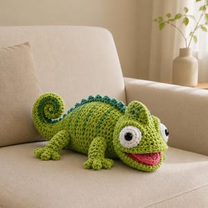 Puede incluir: Un juguete de peluche de camaleón tejido a ganchillo en tonos verde claro y oscuro. La criatura presenta una cola enroscada, una cresta dorsal texturizada, grandes ojos circulares blancos con pupilas negras y una boca abierta que muestra una lengua rosa.