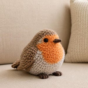 May include: A small, round, crocheted bird figurine featuring a brown back, a bright orange chest, and a light cream-coloured belly. The bird has a dark brown beak and small, black, round eyes, with dark brown crocheted feet at the base.