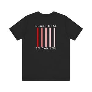 Pode incluir: Camiseta preta com o texto "SCARS HEAL SO CAN YOU" em branco. Há seis listras verticais vermelhas, cor-de-rosa e brancas atrás do texto.
