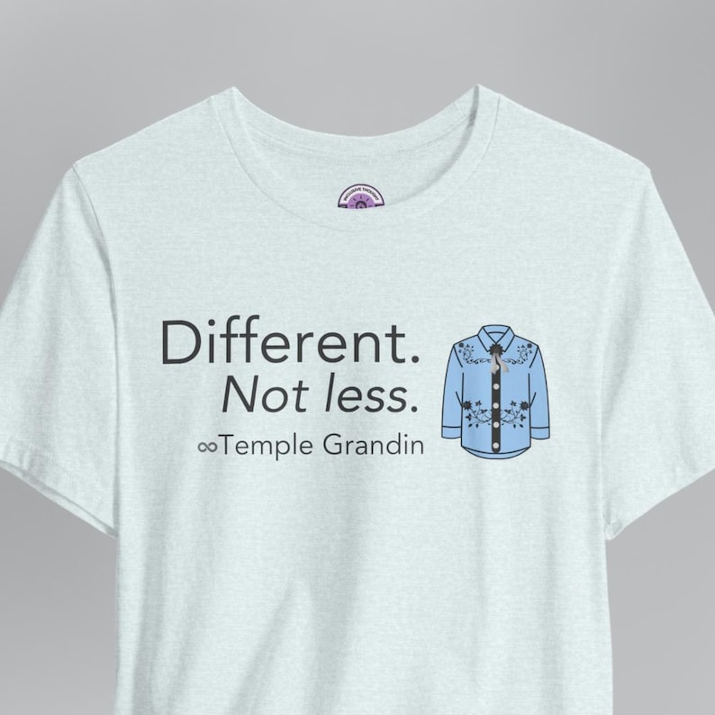 Pu&ograve; includere: Una t-shirt azzurra con il testo nero "Different. Not less." sopra il simbolo dell'infinito e il nome "Temple Grandin". A destra del testo c'&egrave; la grafica di una camicia in stile western azzurra con ricami floreali scuri.