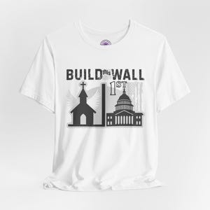 May include: White t-shirt with a graphic design featuring a church, the U.S. Capitol building, and the text "BUILD this WALL 1st". The design incorporates an American flag and angel wings.