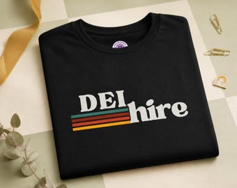 May include: A black short-sleeved t-shirt featuring the white text "DEI hire" printed on the chest. Below the text are four horizontal stripes in teal, dark red, orange, and yellow. The shirt has a round neckline and a simple, clean design.