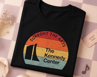 Può includere: Una t-shirt nera a girocollo con una grafica circolare a strisce orizzontali arancioni, gialle e azzurre. Il testo recita SUPPORT THE ARTS e The Kennedy Center, accompagnato da una silhouette nera dell'architettura dell'edificio.