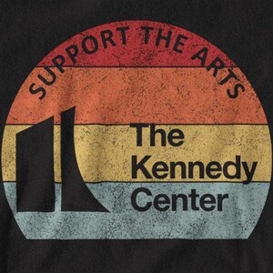 K&ouml;nnte beinhalten: Ein schwarzes T-Shirt mit einer kreisf&ouml;rmigen Grafik aus horizontalen Streifen in Rot, Orange, Gelb und Hellblau. Der Text lautet SUPPORT THE ARTS und The Kennedy Center, erg&auml;nzt durch eine schwarze Silhouette des Kennedy Center Geb&auml;udes.