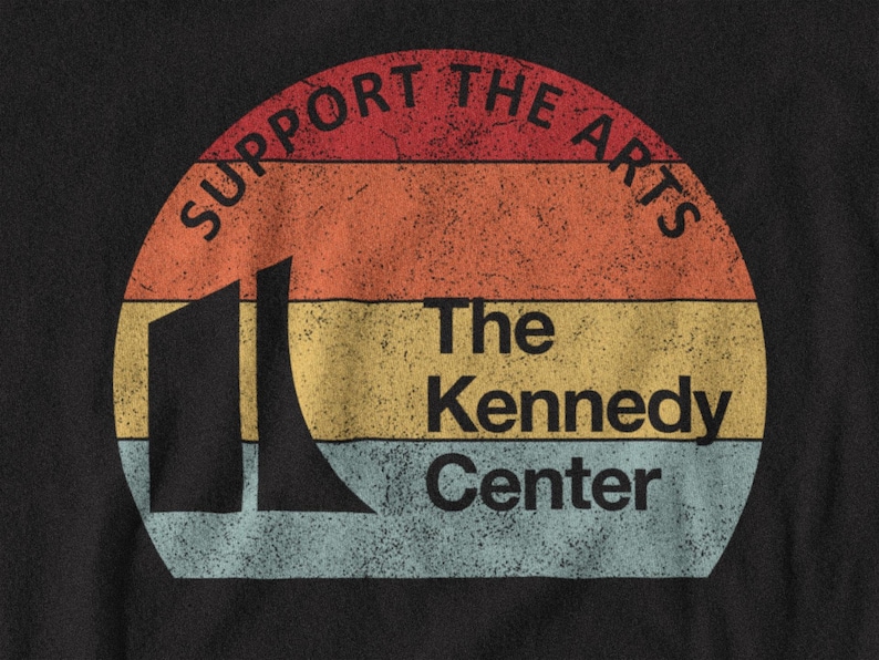 K&ouml;nnte beinhalten: Ein schwarzes T-Shirt mit einer kreisf&ouml;rmigen Grafik aus horizontalen Streifen in Rot, Orange, Gelb und Hellblau. Der Text lautet SUPPORT THE ARTS und The Kennedy Center, erg&auml;nzt durch eine schwarze Silhouette des Kennedy Center Geb&auml;udes.