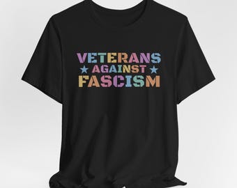 May include: Black t-shirt with the text "VETERANS AGAINST FASCISM" in pastel colors. The text is arranged in three lines with a star on each side of the word "AGAINST".