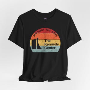 May include: Black t-shirt featuring a retro-style graphic. The design includes a colorful sunset with the text "SUPPORT THE ARTS" arched above a building silhouette and the words "The Kennedy Center" below.