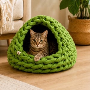 Puede incluir: Una cama para mascotas en forma de cúpula hecha de hilo grueso de punto color verde lima. La estructura presenta una gran abertura circular en la parte frontal, revelando a un gato atigrado marrón descansando dentro. Una pequeña etiqueta beige con una huella está unida al lateral.