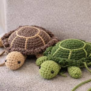 Puede incluir: Dos bolsas de ganchillo con forma de tortuga. Una es marrón con detalles beige, la otra es verde con detalles verde claro. Ambas tienen pequeños ojos negros y una correa. Las tortugas están sobre una superficie gris.