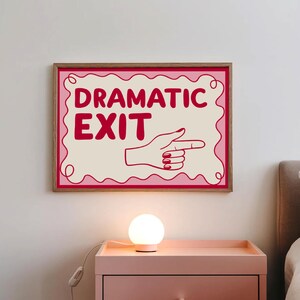 May include: A framed wall art print with the words "DRAMATIC EXIT" in red, with a hand pointing right. The artwork has a cream background with a pink border and is set in a wooden frame. A small, round, white lamp sits on a pink nightstand.