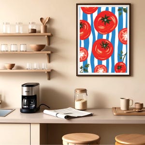 May include: A framed print of red tomatoes on a blue and white striped background. The tomatoes are arranged in a random pattern and some are cut in half. The print is hanging on a light beige wall above a kitchen counter.