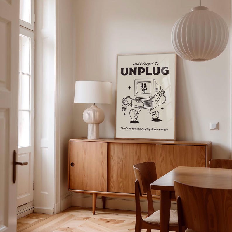 Op de afbeelding: Een ingelijste poster met de tekst "Don't Forget To UNPLUG" en een cartoon computerpersonage. De poster wordt weergegeven op een houten kast, met een witte lamp en een ronde armatuur op de achtergrond. De afbeelding heeft een vintage esthetiek.