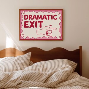 May include: A framed wall art print with the words "DRAMATIC EXIT" in red letters. The print features a hand pointing to the right. The frame is brown, and the background is a light beige color. The art is above a bed.