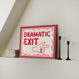 May include: A framed art print with the words "DRAMATIC EXIT" in red, featuring a hand pointing right. The print has a pink border and is set in a wooden frame. Two black candle holders are to the right.