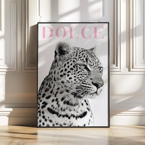 May include: Black framed art print featuring a black and white leopard portrait. The word "DOLCE" is printed in pink at the top. The artwork is displayed on a white wall and a wooden floor.