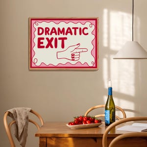 May include: A framed wall art print with the words "DRAMATIC EXIT" in red, along with a hand pointing to the right. The print has a pink border and is set against a cream background. The art is displayed above a wooden table.