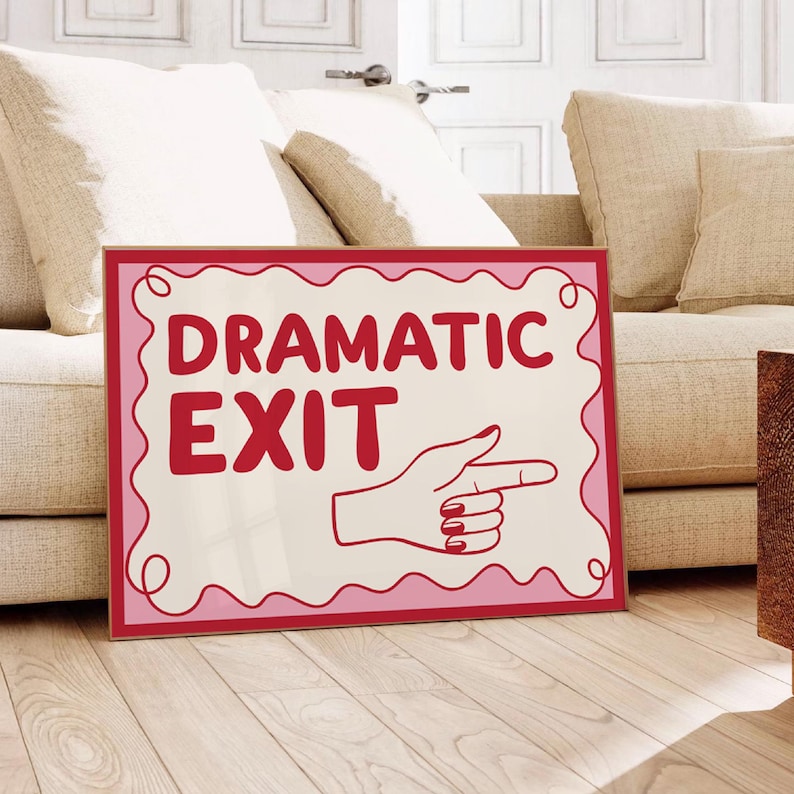 May include: A framed sign with the words "DRAMATIC EXIT" in red, with a pointing hand graphic. The sign has a pink and red border and is displayed on a wooden floor.