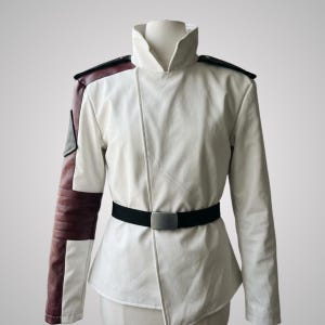 May include: A white jacket featuring a high collar, long sleeves, and an asymmetrical front closure. The right sleeve includes dark maroon panels and a triangular patch. A black belt with a rectangular silver buckle is worn around the waist.