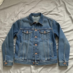 May include: A blue denim jacket with copper-coloured buttons and two chest pockets. The jacket features a collared neckline and long sleeves. The denim has a medium wash and a slightly worn appearance.