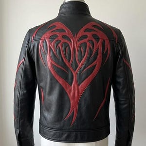 May include: Black leather jacket featuring a red heart-shaped design on the back. The jacket has a stand-up collar and a zip closure. The red design is in a tribal style.