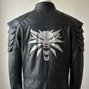 May include: Black leather jacket featuring a white and red wolf emblem on the back. The jacket has shoulder armor detailing and a stand-up collar. The wolf emblem is a stylized design with sharp angles and red eyes, adding a bold statement to the jacket's design.