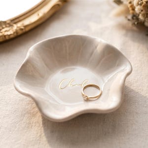 Pu&ograve; includere: Un piccolo piatto portagioie in ceramica bianco sporco con un bordo ondulato e la scritta "Chand" in oro. Un anello d'oro con un piccolo diamante &egrave; all'interno del piatto. Il piatto &egrave; su una superficie di colore neutro.