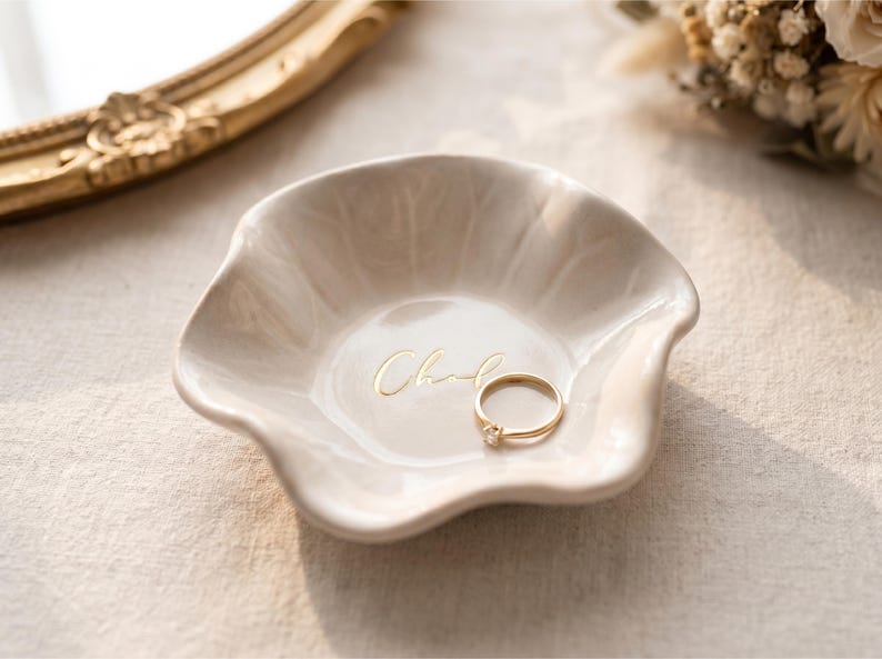 Pu&ograve; includere: Un piccolo piatto portagioie in ceramica bianco sporco con un bordo ondulato e la scritta "Chand" in oro. Un anello d'oro con un piccolo diamante &egrave; all'interno del piatto. Il piatto &egrave; su una superficie di colore neutro.