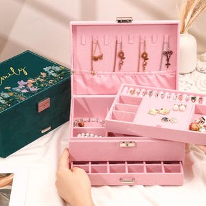 Può includere: Un portagioie rosa con più scomparti e un vassoio estraibile, pieno di varie collane, anelli e orecchini. È visibile anche un secondo portagioie, color verde acqua, con decorazioni floreali e il nome "Emily" in oro.