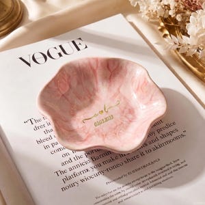 Pu&ograve; includere: Un portagioie rosa a forma di petalo con un motivo marmorizzato. Il piatto ha scritte dorate con le iniziali "A & L" e la data "02.03.2023". Il piatto &egrave; su una rivista Vogue.