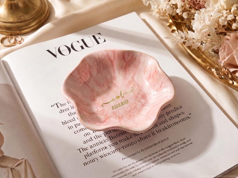 Pu&ograve; includere: Un portagioie rosa a forma di petalo con un motivo marmorizzato. Il piatto ha scritte dorate con le iniziali "A & L" e la data "02.03.2023". Il piatto &egrave; su una rivista Vogue.